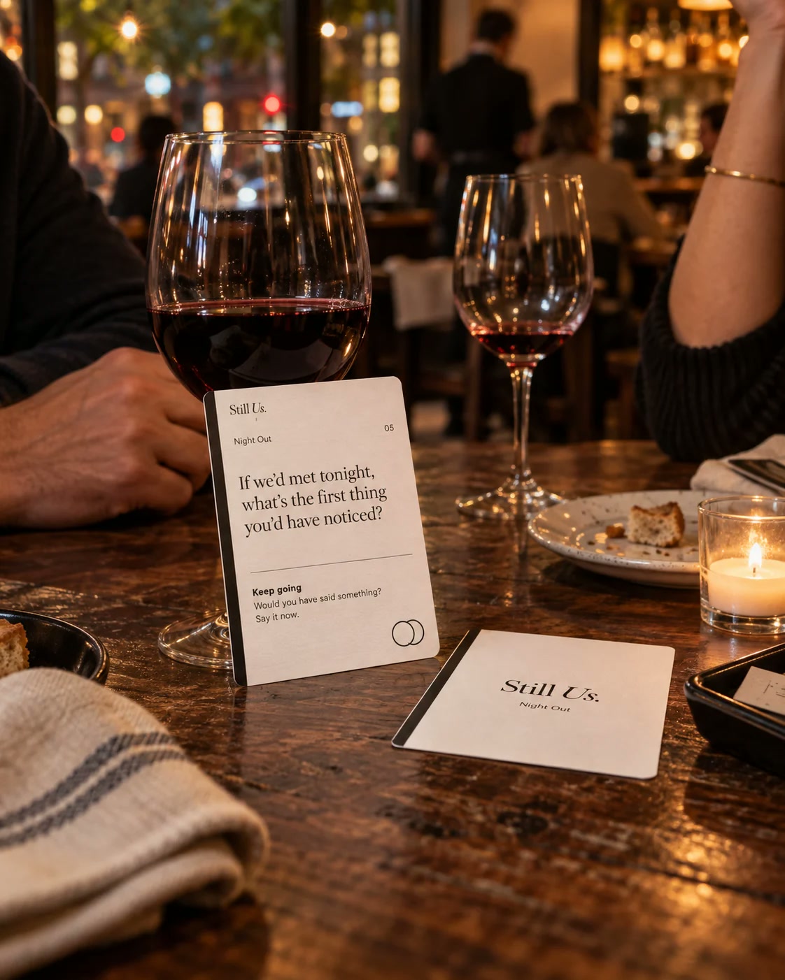 A Still Us Night Out card standing against a glass of red wine on a restaurant table, a second card face-down beside it.